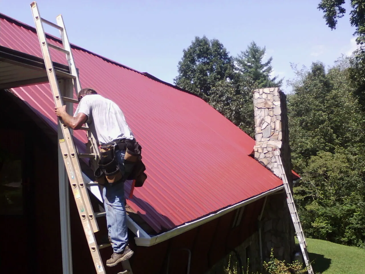 Roofer on a ladder beside a red metal roof and stone chimney — Soffit & Fascia Repair by Roofing Brothers in Woodland Hills